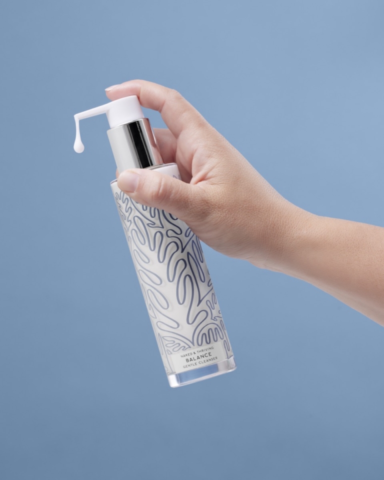 Woman's hand holding Naked & Thriving Balance Gentle Cleanser pump bottle against a blue background with a single drop of white cleanser visible on the pump nozzle, showing product dispensing in use
