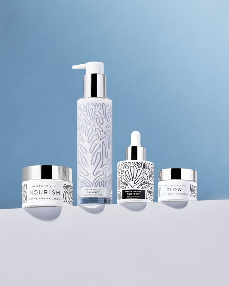 Four Naked & Thriving skincare products arranged on a blue gradient surface: Nourish Active Barrier Cream jar, Balance Gentle Cleanser pump bottle, Protect Face Serum dropper, and Glow Triple Boost Eye Cream jar, all featuring white and silver packaging with organic botanical line art