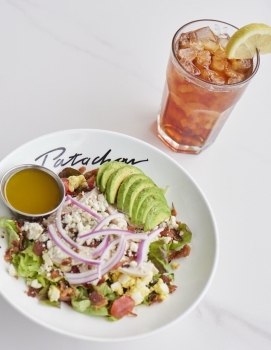 Patachou Cobb salad with sliced avocado, red onion, crumbled blue cheese, bacon, egg, and mixed greens in a branded Patachou bowl with a side of vinaigrette, served alongside an iced tea with lemon on a white marble surface