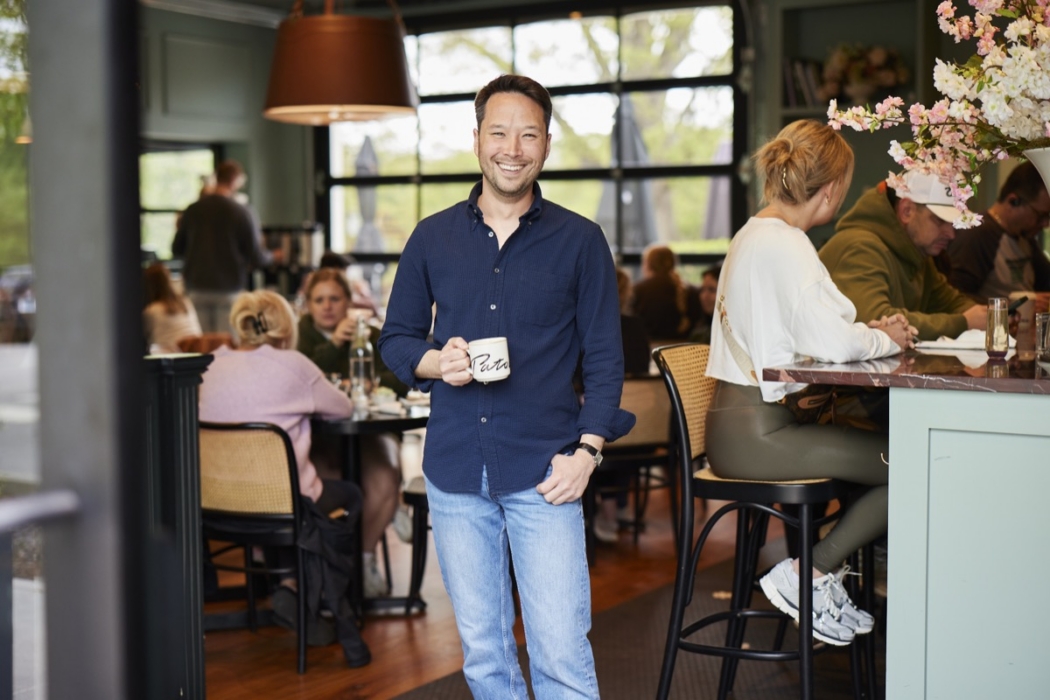 Smiling man in navy blue shirt holding a Patachou branded coffee mug, standing in a bright, fully-seated Café Patachou dining room with cherry blossom floral arrangement and warm natural light through large windows