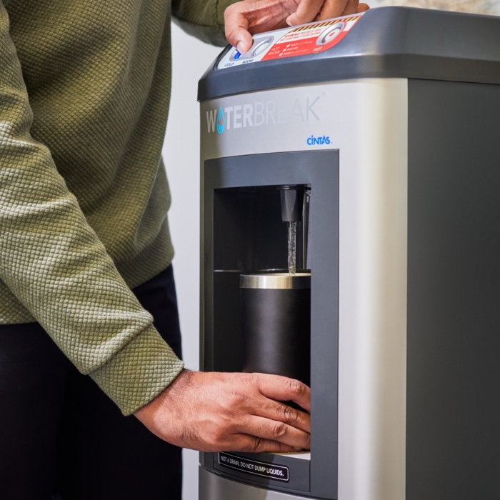 Person filling a black insulated tumbler at a Cintas WaterBreak bottleless water cooler dispenser, pressing the cold water button, in an office or workplace break room setting