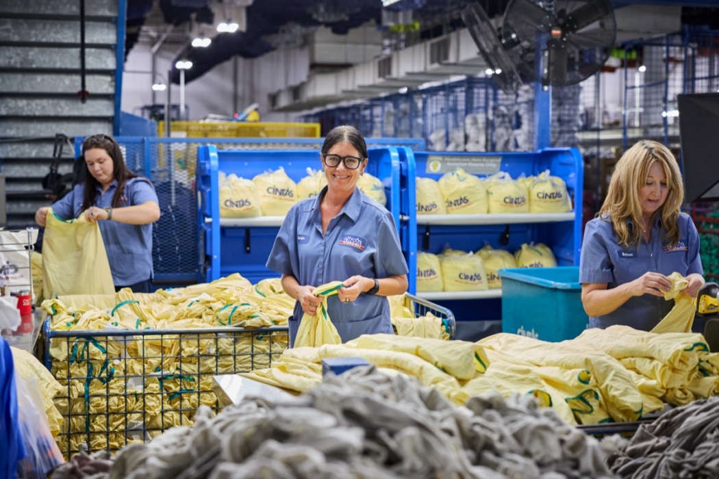Three smiling Cintas employees in blue uniforms sorting yellow garment bags on a processing floor inside a large commercial laundry and uniform services facility