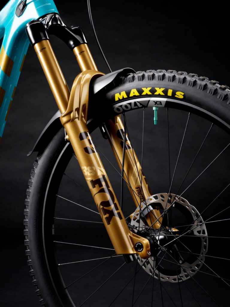 Close-up of teal Yeti mountain bike front fork with gold Fox suspension, Maxxis tire, and disc brake rotor against black background