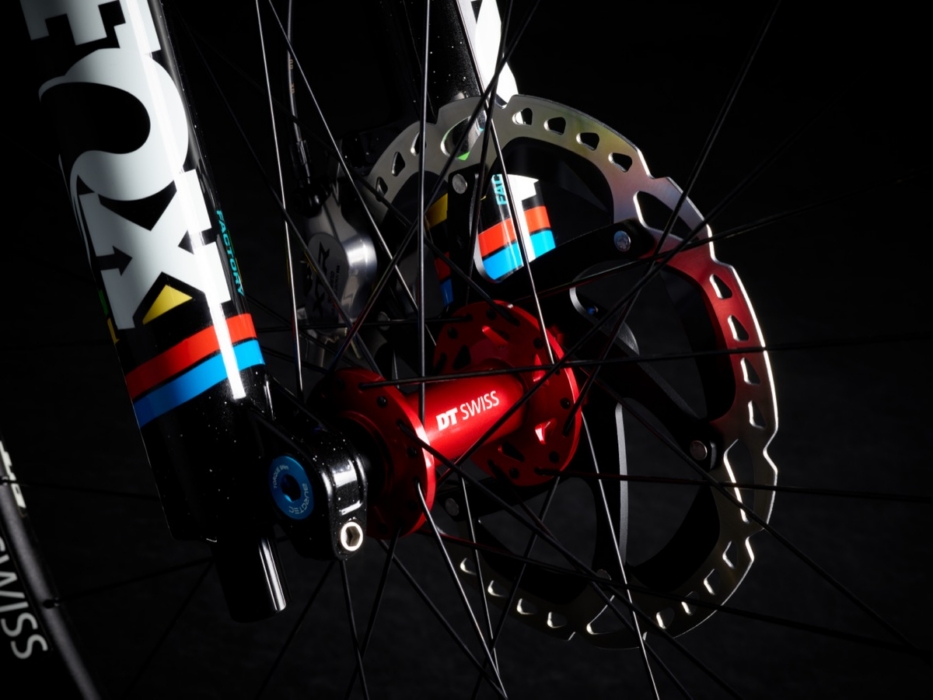 Macro studio photo of red DT Swiss front hub and disc brake rotor on Yeti SB160 mountain bike, with Fox 40 Factory fork leg showing UCI World Champion rainbow stripes and black spoke web in dramatic low-key lighting