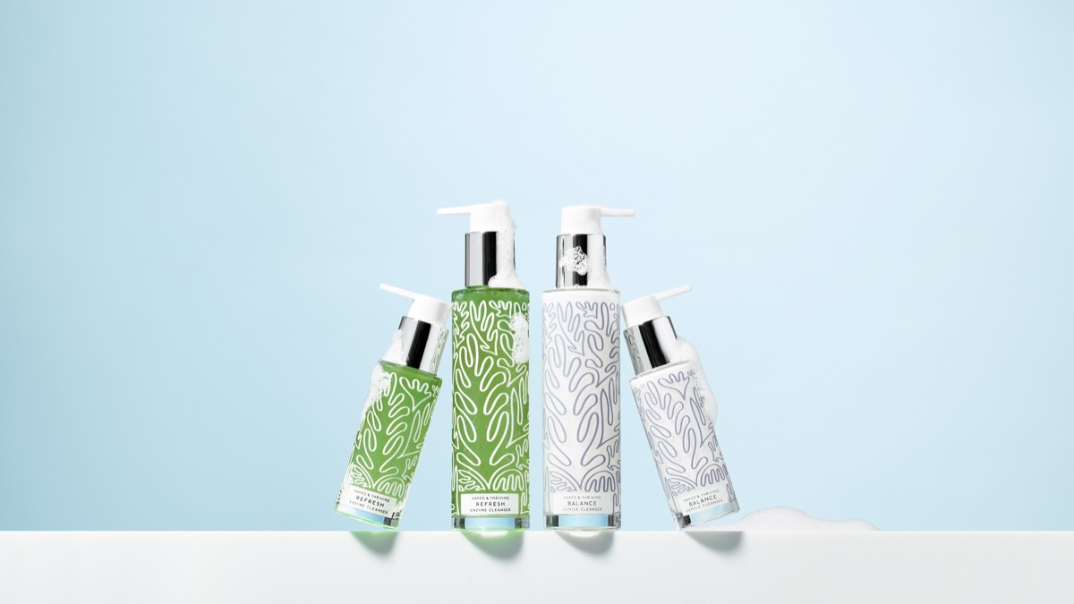 Four bottles of Naked and Thriving Refresh Enzyme Cleanser and Balance Gentle Cleanser with foam texture on white ledge against blue background