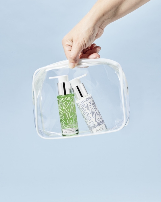 Hand holding a clear travel bag containing Naked and Thriving Refresh Enzyme Cleanser and Balance Gentle Cleanser against light blue background
