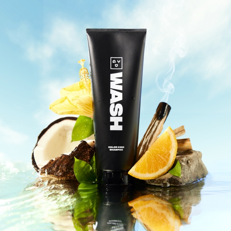 Black tube of GD Wash Color Kind Shampoo standing on rocks with coconut, orange slice, hibiscus flower, and smoking palo santo against sky backdrop