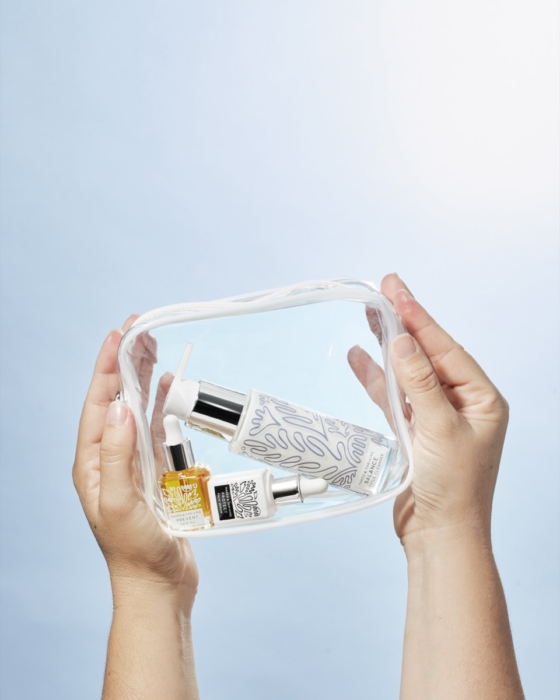 Two hands holding a clear travel pouch containing Naked and Thriving Balance Cleanser, Protect Face Serum, and Prevent Face Oil against light blue background