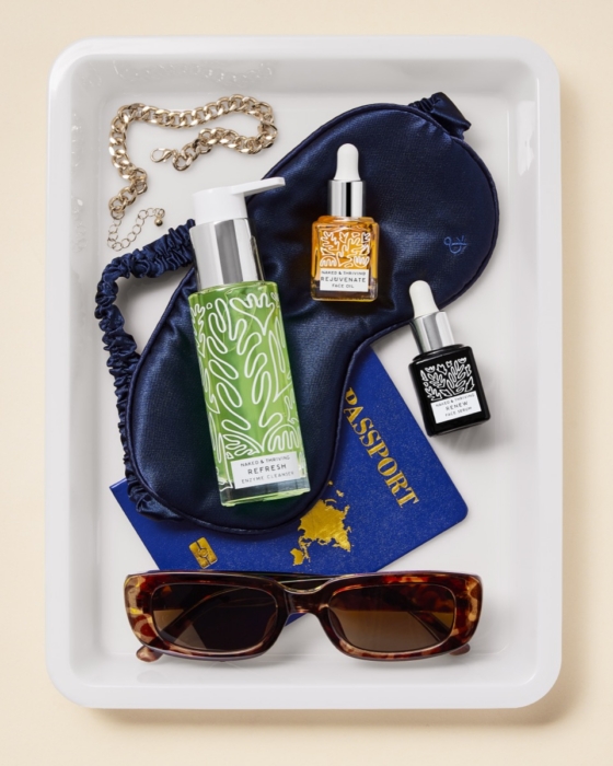 Naked and Thriving Refresh Enzyme Cleanser, Rejuvenate Face Oil, and Renew Face Serum styled on white tray with sleep mask, gold jewelry, passport, and sunglasses