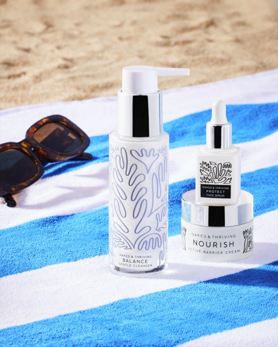Naked and Thriving Balance Cleanser, Protect Face Serum, and Nourish Active Barrier Cream styled on blue striped beach towel with sunglasses and sand