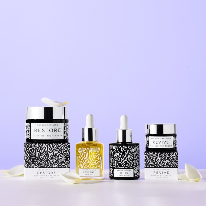 Naked & Thriving full skincare collection bottles and jars flat lay with white florals on lavender background