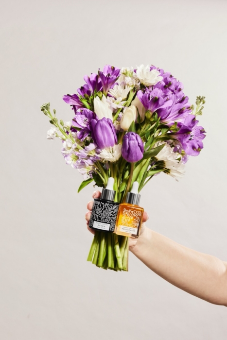 Hand holding purple tulip bouquet with Naked & Thriving facial oil bottle on white background