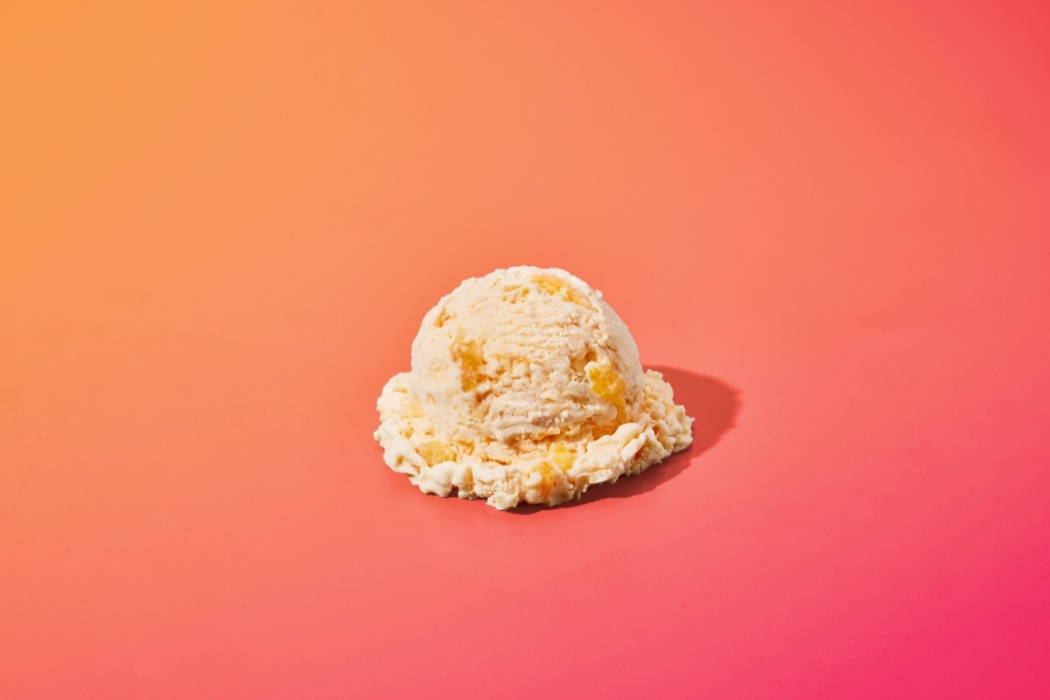 Single scoop of peach ice cream with visible fruit chunks on colorful orange to pink gradient background