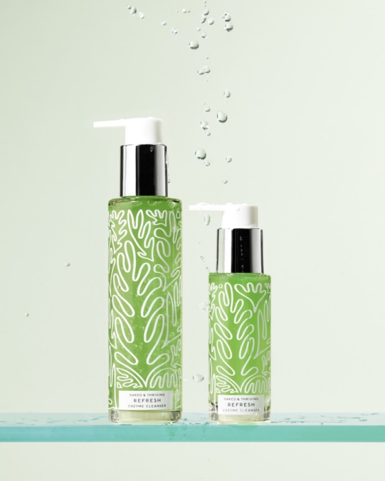 Two sizes of Naked and Thriving Refresh Enzyme Cleanser green pump bottles with white leaf pattern on glass shelf with water droplets