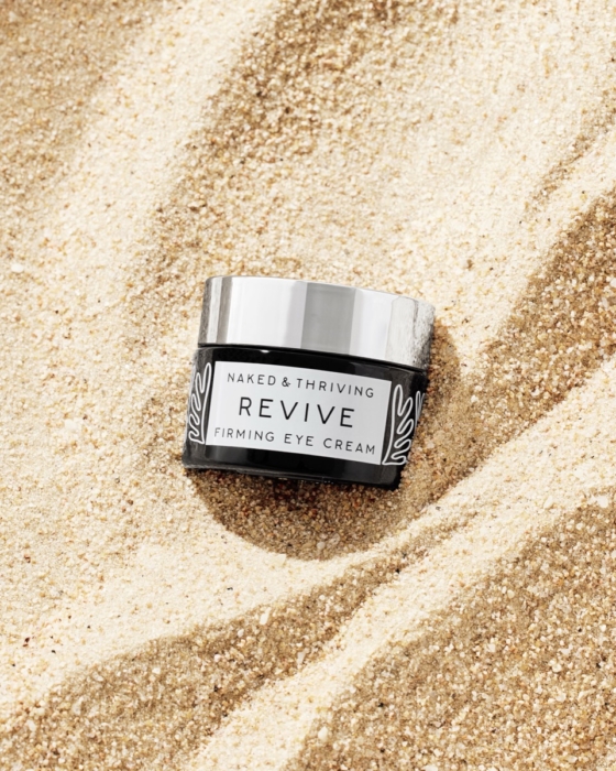 Naked and Thriving Revive Firming Eye Cream jar with silver lid nestled in swirled beach sand