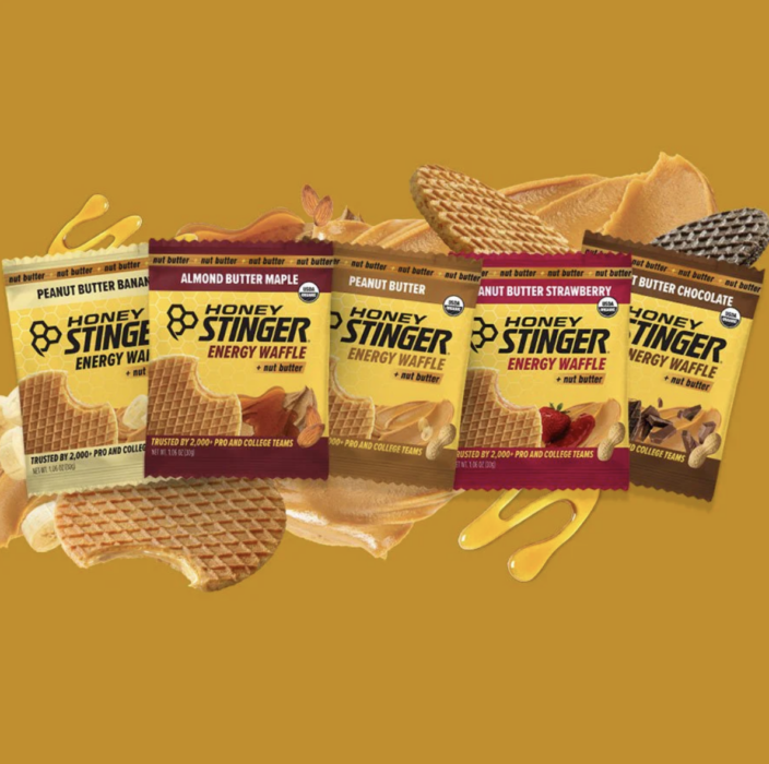 Five Honey Stinger Energy Waffle nut butter flavor pouches including peanut butter banana, almond butter maple, peanut butter, peanut butter strawberry, and peanut butter chocolate with waffle and fruit props