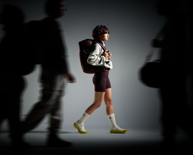 Woman in adidas cycling shorts and cream jacket walking with maroon backpack, framed by blurred silhouettes in dramatic studio lighting