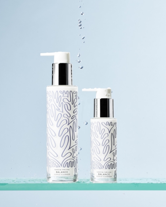 Two sizes of Naked and Thriving Balance Gentle Cleanser white pump bottles with leaf pattern on glass shelf with water droplets