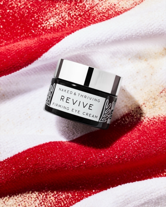 Naked and Thriving Revive Firming Eye Cream jar with silver lid resting on red and white striped towel with scattered sand