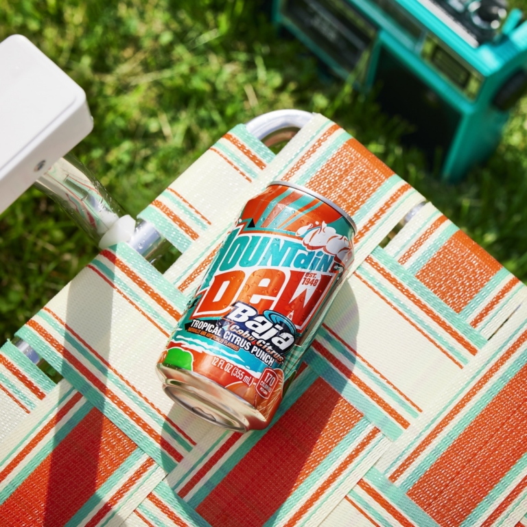 Can of Mountain Dew Baja Blast resting on a striped orange and teal lounge chair beside a sprinkler and green lawn furniture