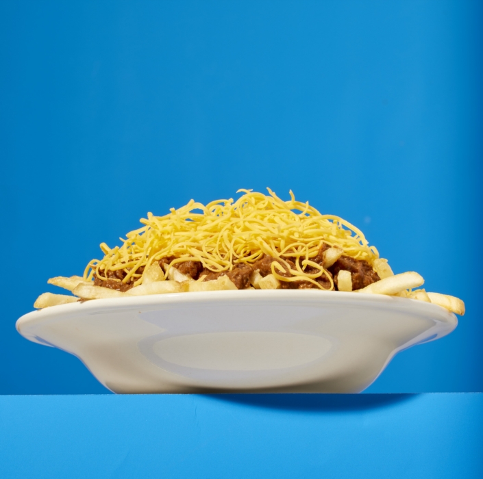 Side view of a bowl piled high with Cincinnati style chili, fries, and shredded cheese against a solid blue background