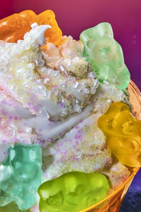 Close-up of vanilla soft serve topped with colorful gummy bears and sanding sugar against a pink and purple gradient background