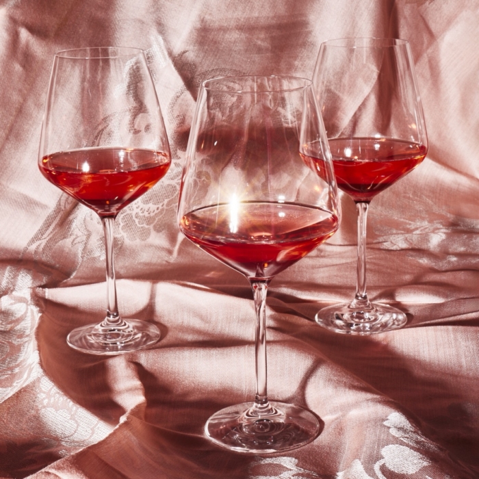 Three crystal wine glasses filled with red wine arranged on shimmering pink satin fabric with candlelight reflection
