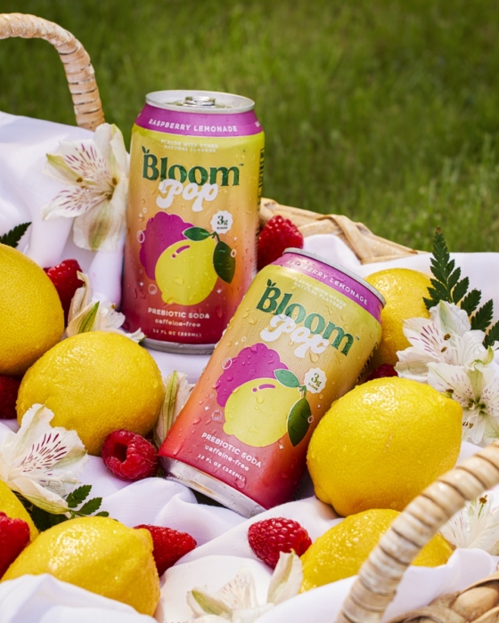 Two cans of Bloom Pop sparkling raspberry lemonade in a woven basket surrounded by fresh lemons, raspberries, and white flowers