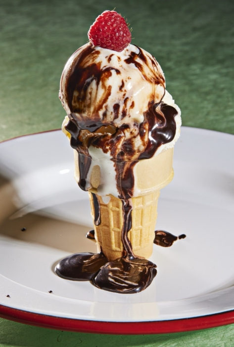 Waffle cone with a scoop of vanilla ice cream drizzled in chocolate sauce and topped with a fresh raspberry, dripping onto a white plate with red rim