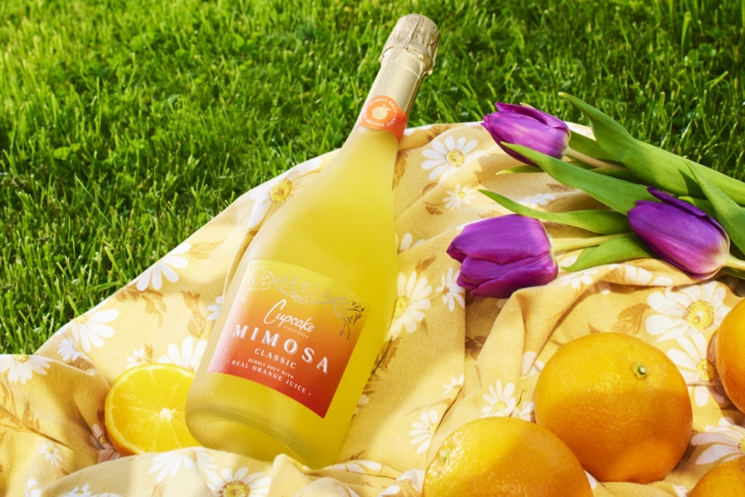 Bottle of Corinne's Concoctions Mimosa cocktail lying on a floral picnic blanket with purple tulips and fresh oranges on green grass