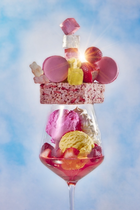 Elaborate dessert wine glass filled with pink and yellow ice cream, strawberries, and topped with macarons, gummy bears, and a rice cereal treat crown against a cloudy sky background