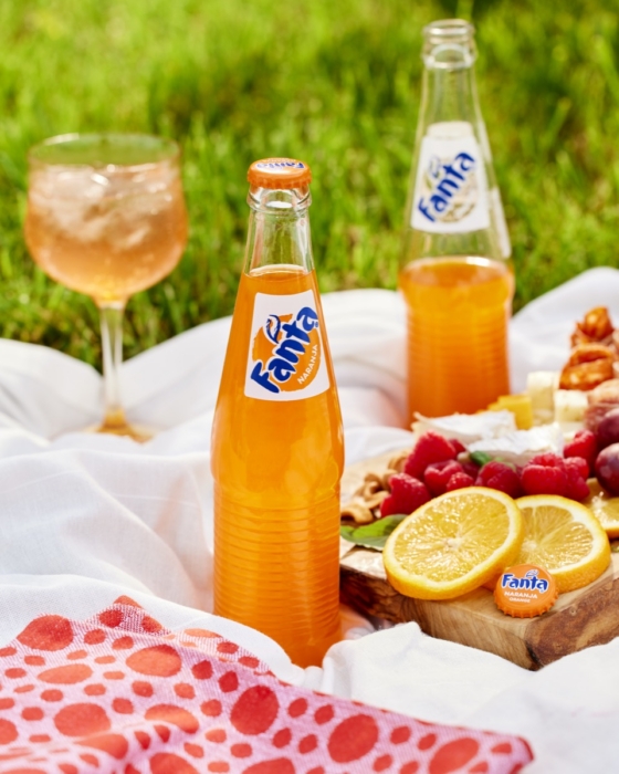 Two glass bottles of Fanta orange soda on a white picnic blanket with a poured glass, sliced lemons, and a charcuterie board with berries