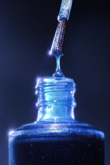 Glittery blue nail polish brush dripping a bead of polish back into an open glass bottle against a dark background