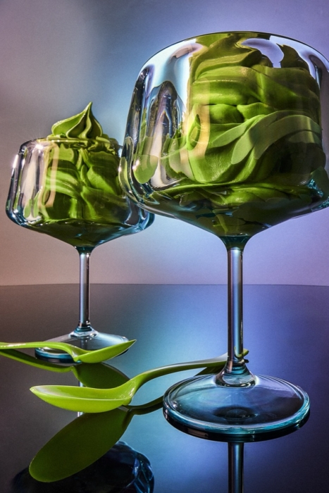 Two stemmed glasses filled with swirled green soft serve ice cream reflected dramatically against a purple and blue gradient background