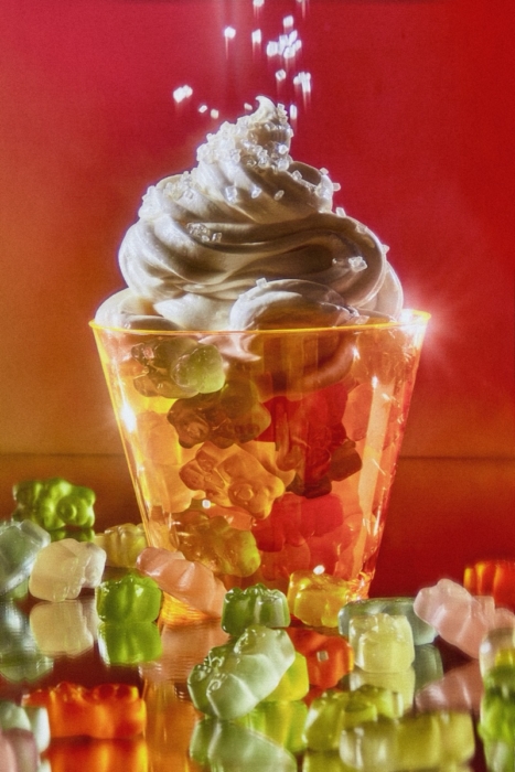Glass cup filled with colorful gummy bears topped with swirled soft serve and sanding sugar, surrounded by scattered gummy bears on a red background