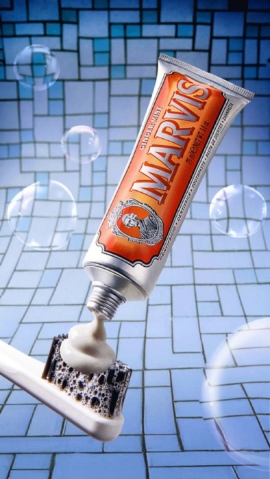 Marvis ginger mint toothpaste tube squeezing paste onto a black toothbrush against a blue tiled bathroom background with bubbles