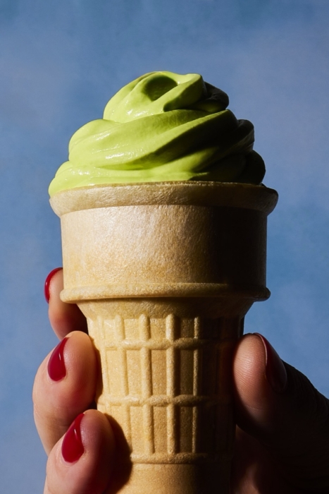 Close-up of a hand with red nail polish holding a waffle cone topped with swirled green soft serve ice cream against a blue sky background