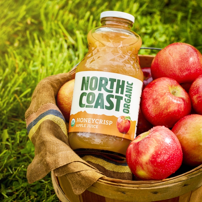Bottle of North Coast Organic Honeycrisp apple juice nestled in a wooden basket filled with fresh red apples on green grass