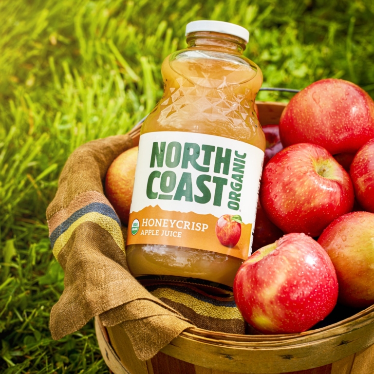 Bottle of North Coast Organic Honeycrisp apple juice nestled in a wooden basket filled with fresh red apples on green grass