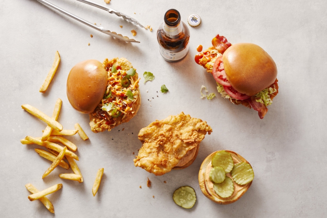 Overhead flat lay of fried chicken sandwiches, loose fries, and a beer bottle arranged on a light gray surface