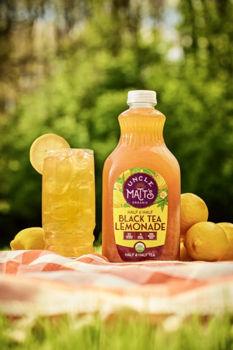 Bottle of Uncle Matt's Organic Half & Half Black Tea Lemonade beside a glass of iced tea lemonade with a lemon wheel garnish, surrounded by fresh lemons on a picnic blanket outdoors
