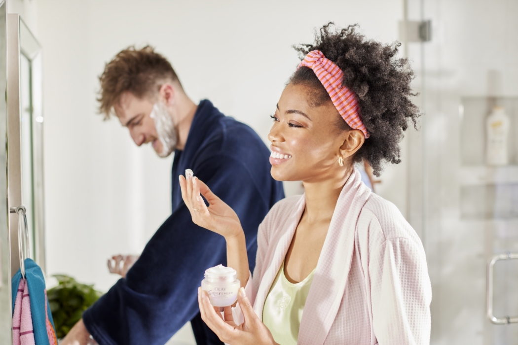 Smiling woman with a pink headband applying cream to her face while a man dances playfully in the background
