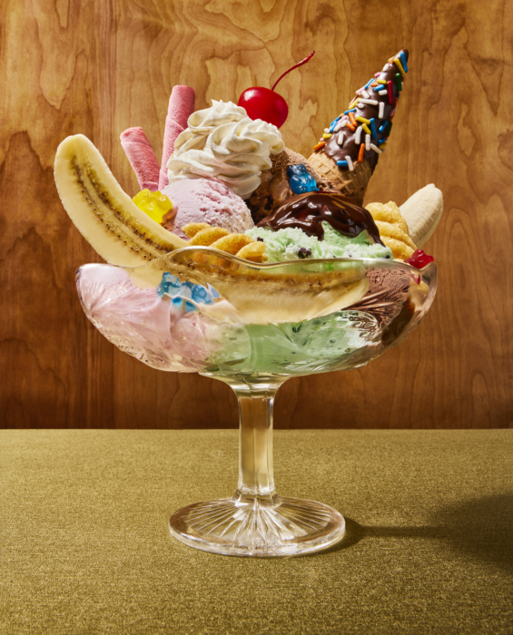 Banana split sundae in a glass pedestal dish with pink, chocolate, and mint chip ice cream, whipped cream, chocolate drizzle, wafer rolls, a chocolate sprinkle waffle cone, gummy bears, and a maraschino cherry on a gold background