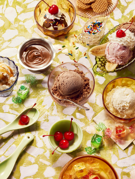 Overhead flat lay of an ice cream sundae party spread with bowls of chocolate and vanilla ice cream, hot fudge, sprinkles, maraschino cherries, banana splits, and wrapped candies on a green marbled surface