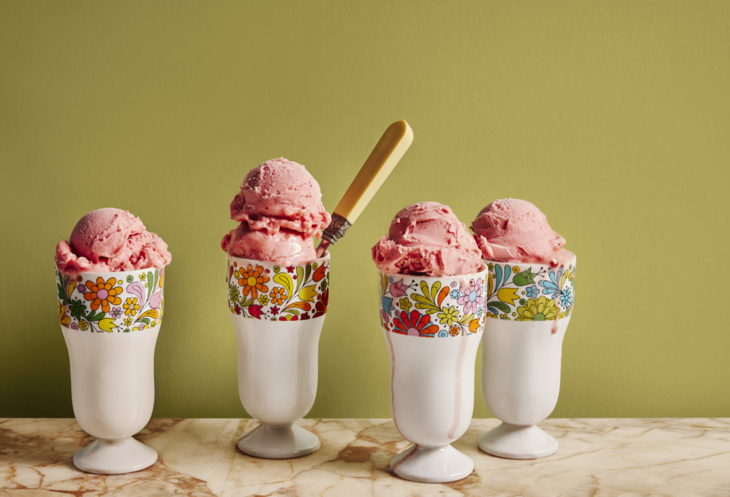 Four footed glass cups with colorful retro floral bands filled with scoops of pink strawberry ice cream and a yellow spoon, on a marble surface with olive green background