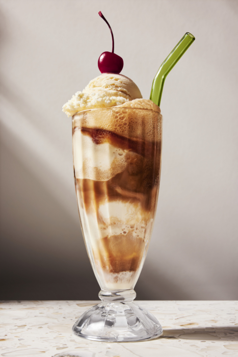 Tall fountain glass root beer float with foamy head, scoop of vanilla ice cream, maraschino cherry, and green glass straw on a marble counter