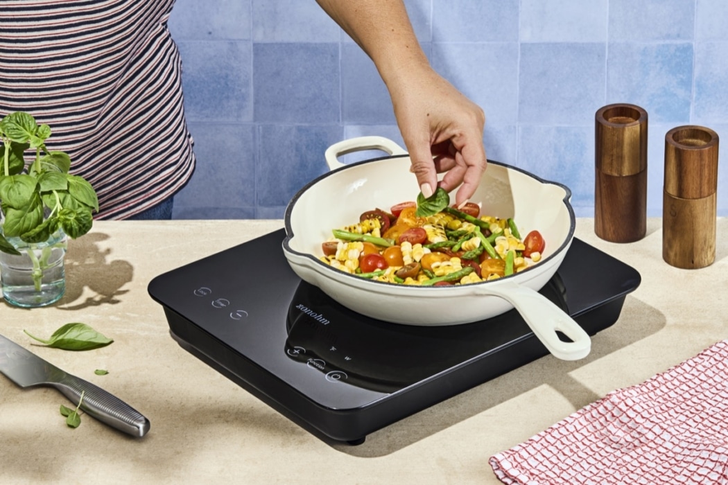 Person adding a fresh basil leaf to a corn, cherry tomato, and asparagus salad in a cream pan on a black Zonohm induction cooktop, styled against a blue tile backdrop
