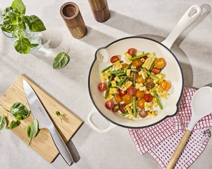 Overhead shot of grilled corn, cherry tomatoes, and asparagus in a cream enameled cast iron pan, styled with fresh basil, wooden pepper mills, and a red-and-white checkered towel