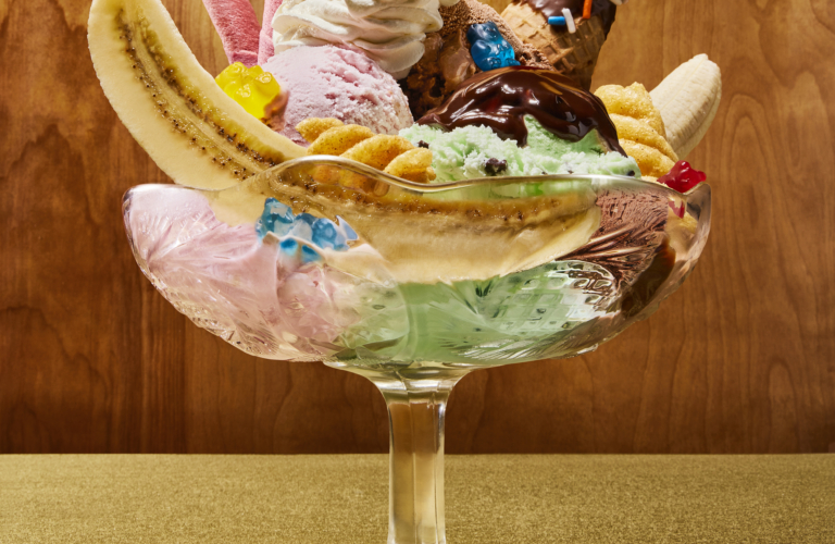 Banana split sundae in a glass pedestal dish with pink, chocolate, and mint chip ice cream, whipped cream, chocolate drizzle, wafer rolls, a chocolate sprinkle waffle cone, gummy bears, and a maraschino cherry on a gold background