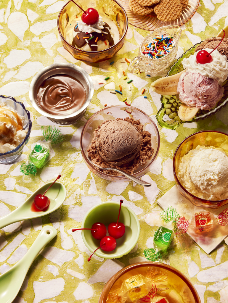 Overhead flat lay of an ice cream sundae party spread with bowls of chocolate and vanilla ice cream, hot fudge, sprinkles, maraschino cherries, banana splits, and wrapped candies on a green marbled surface